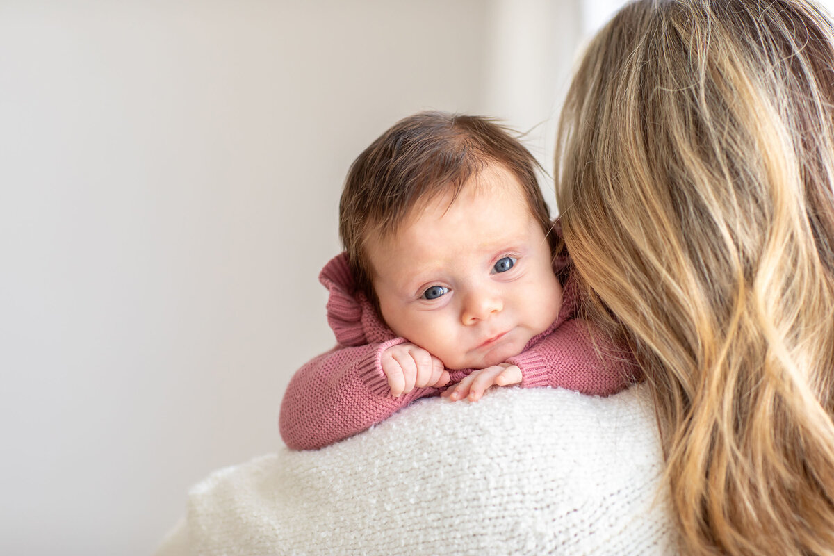 Bucks County family photographers capture a heartwarming moment of a mother holding her baby over her shoulder, with the baby gazing curiously at the camera with wide eyes