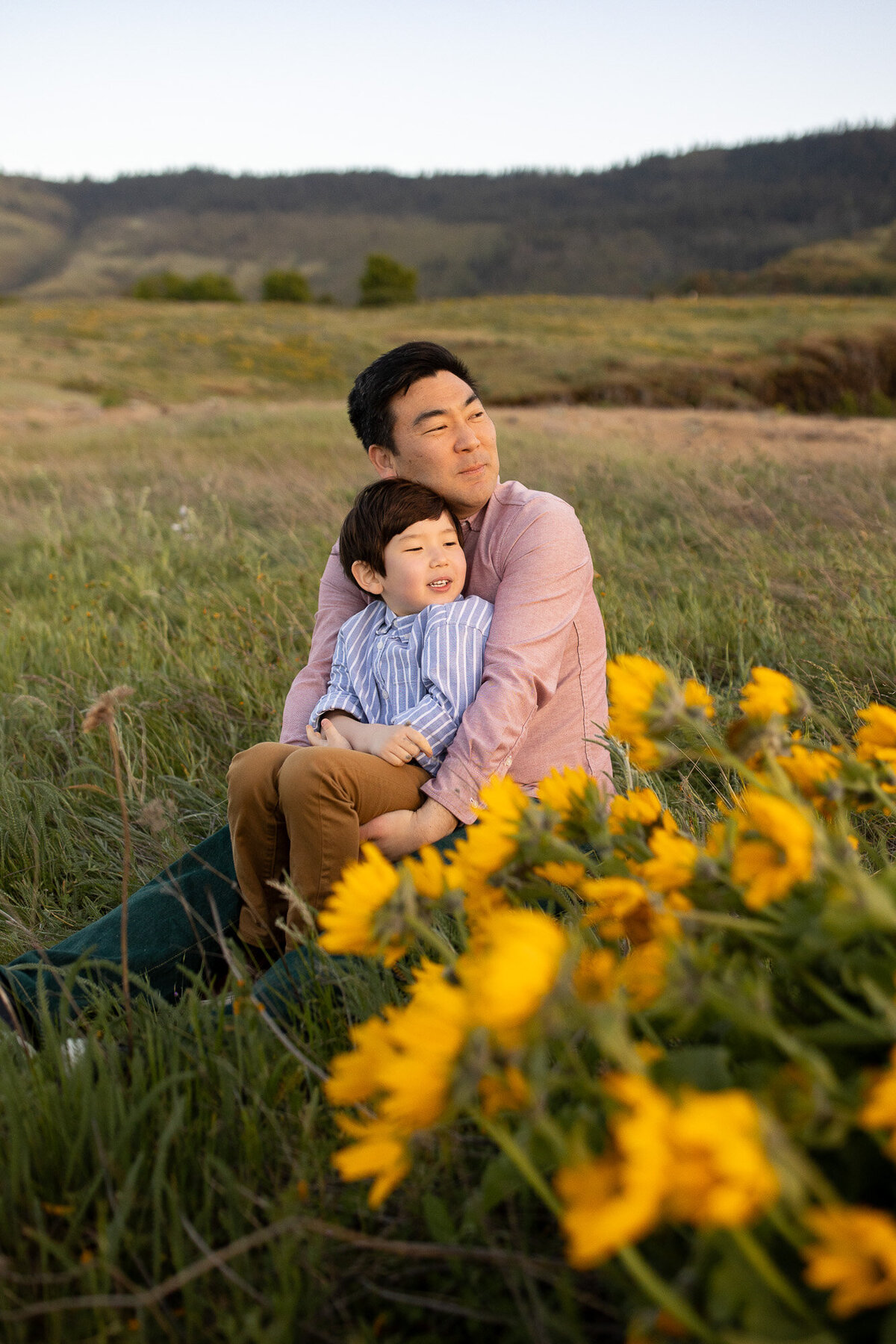 Father and son sitting in tall green grass next to yellow wildflowers in the Gorge at sunset. 