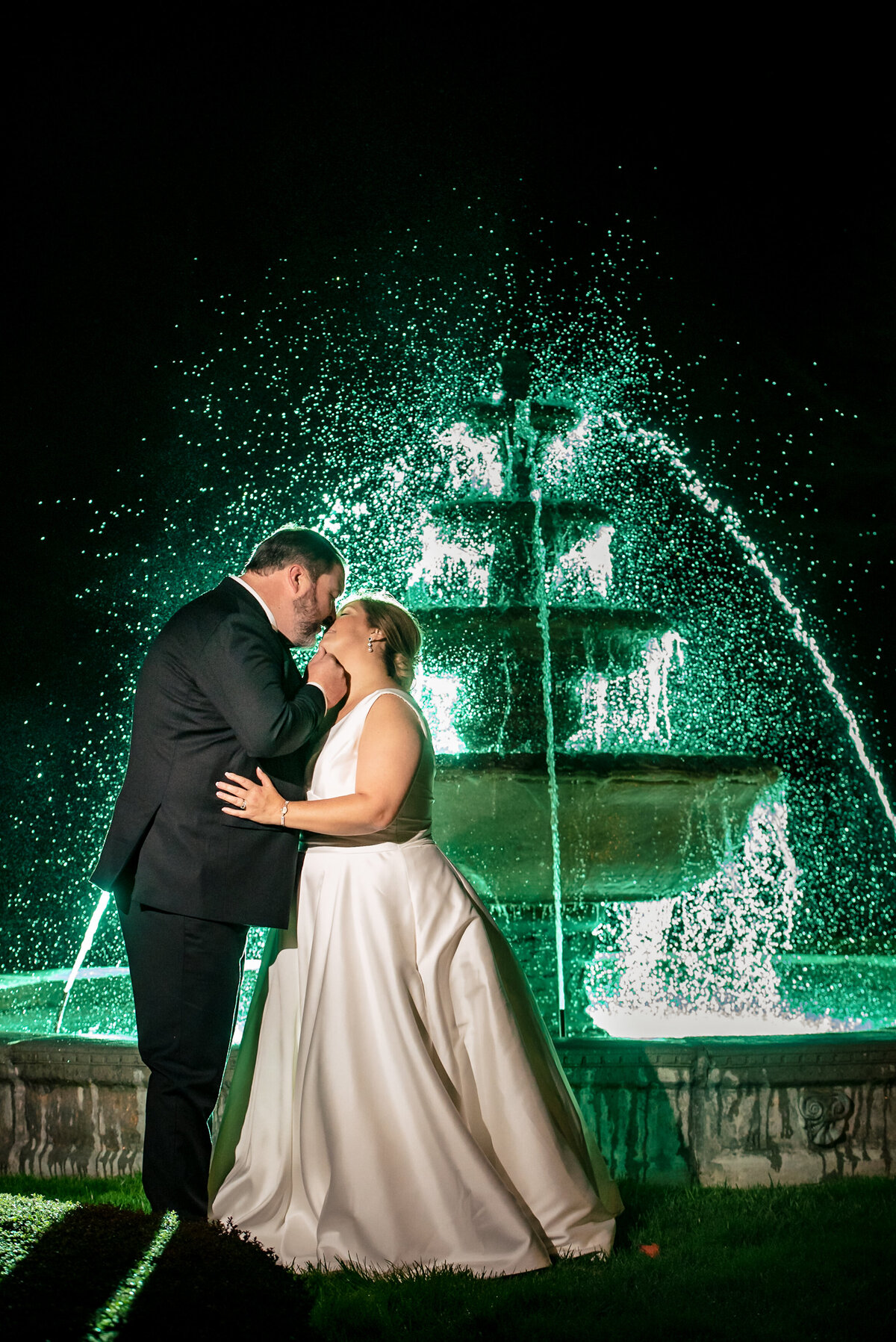 A-nighttime-portrait-of-a-bride-and-groom-kissing-in-front-of-a-lit-up-green-fountain-at-The-Club-at-Longview-in-Charlotte-NC