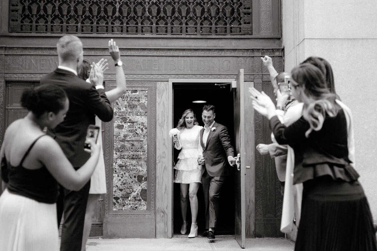 The bride and groom are happily going out of the exit door of NY City Hall after their elopement, as their friends cheer and take pictures.