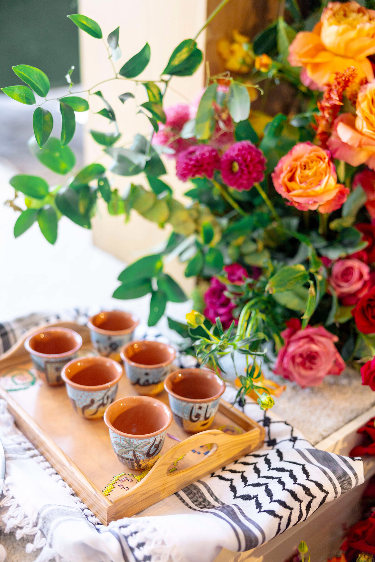A wooden tray with six decorative cups sits atop a black-and-white patterned cloth. The setting is adorned with vibrant flowers in shades of pink, orange, and red, alongside lush green leaves.
