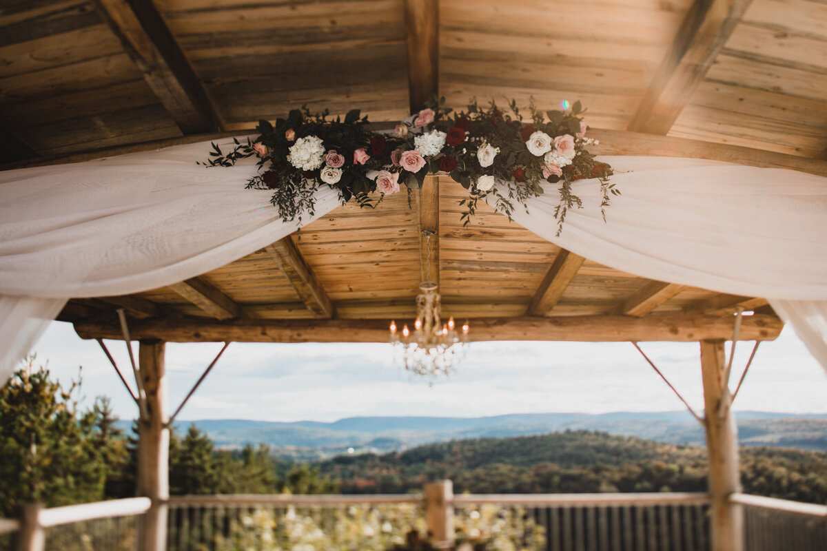 A long burgundy and blush floral installation is placed on a wood beam with white chiffon draping under a large wood canopy with a chandelier as a romantic backdrop for a wedding ceremony at Le Belvedere venue in Wakefield Quebec