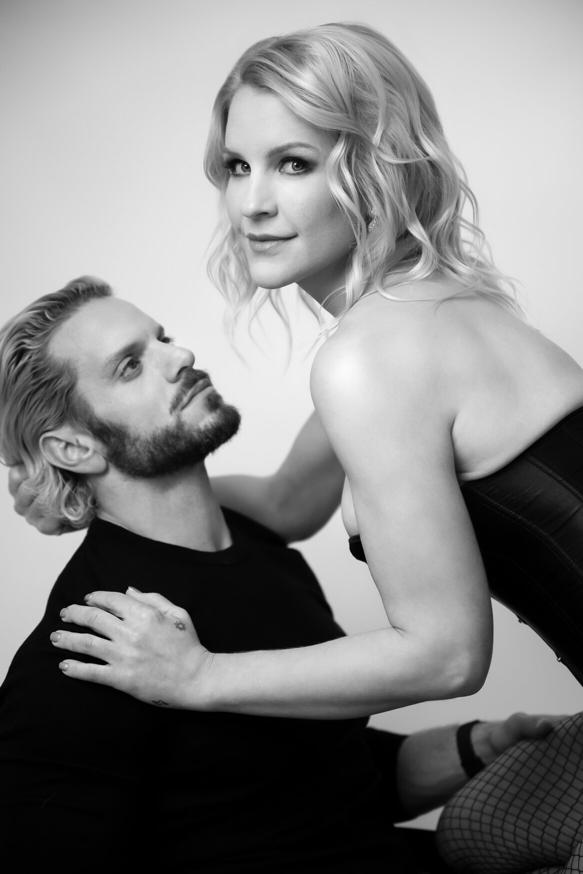 Black and white close-up portrait of a couple in a romantic moment. The woman, wearing a corset and fishnet stockings, looks confidently into the camera with a soft smile, while the man gazes up at her, his hand resting on her waist.