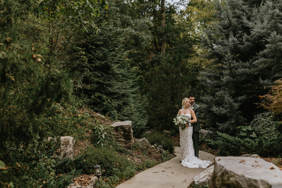 bride and groom stand in pine trees as bride lays back against her husband and he hugs her
