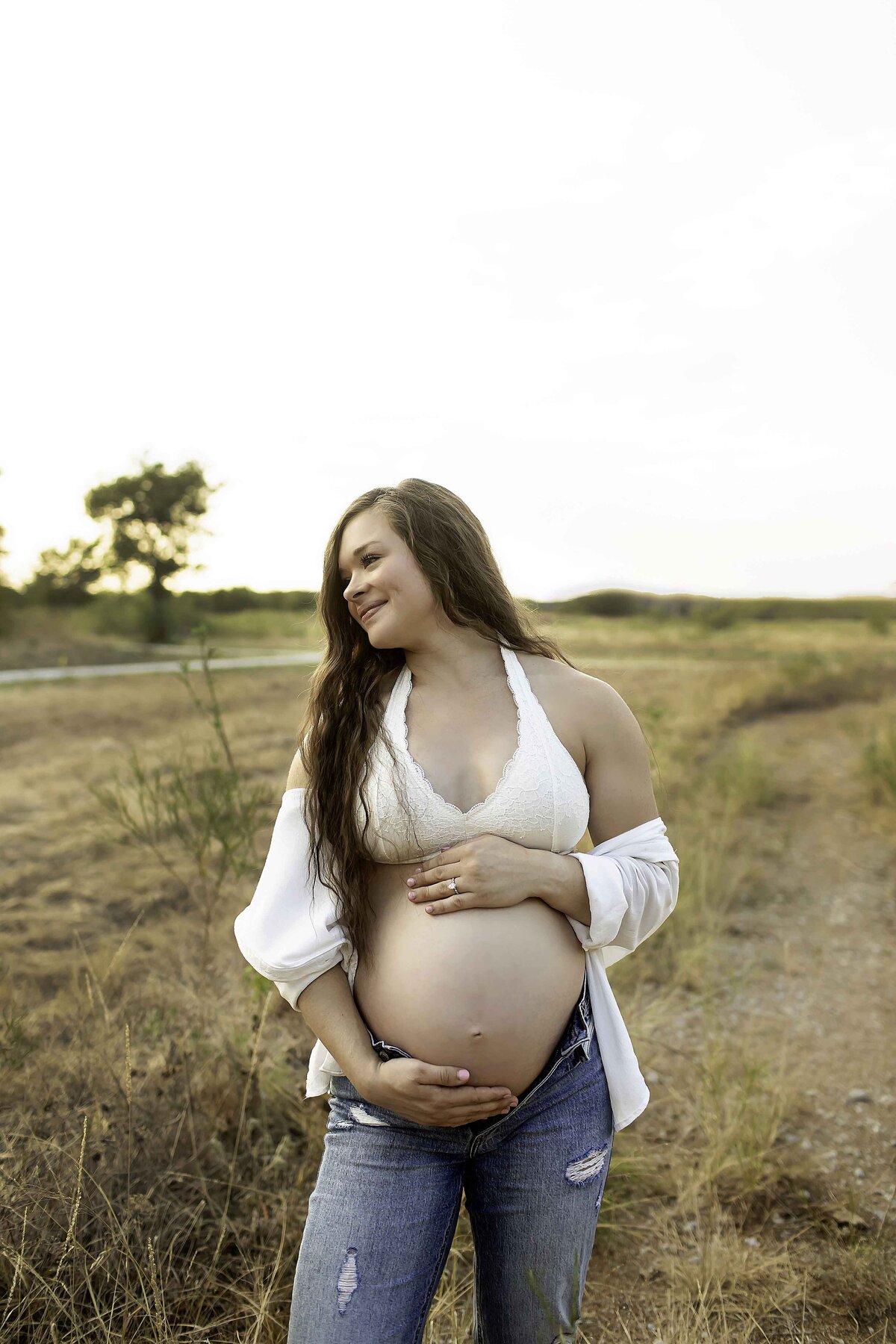 A peaceful maternity photo of the expectant mom standing in a field of white flowers, gently cradling her belly with a soft smile, captured by Chunky Monkey Photography.