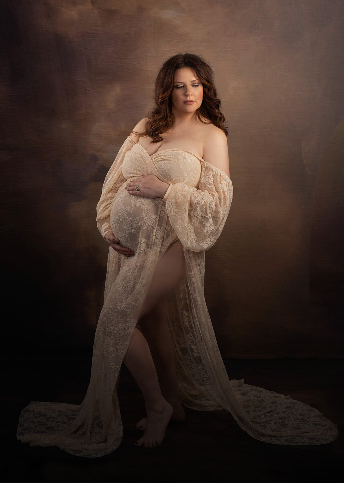 A mother to be with red hair gazes over her shoulder while holding her bump in a cream lace maternity gown in a studio