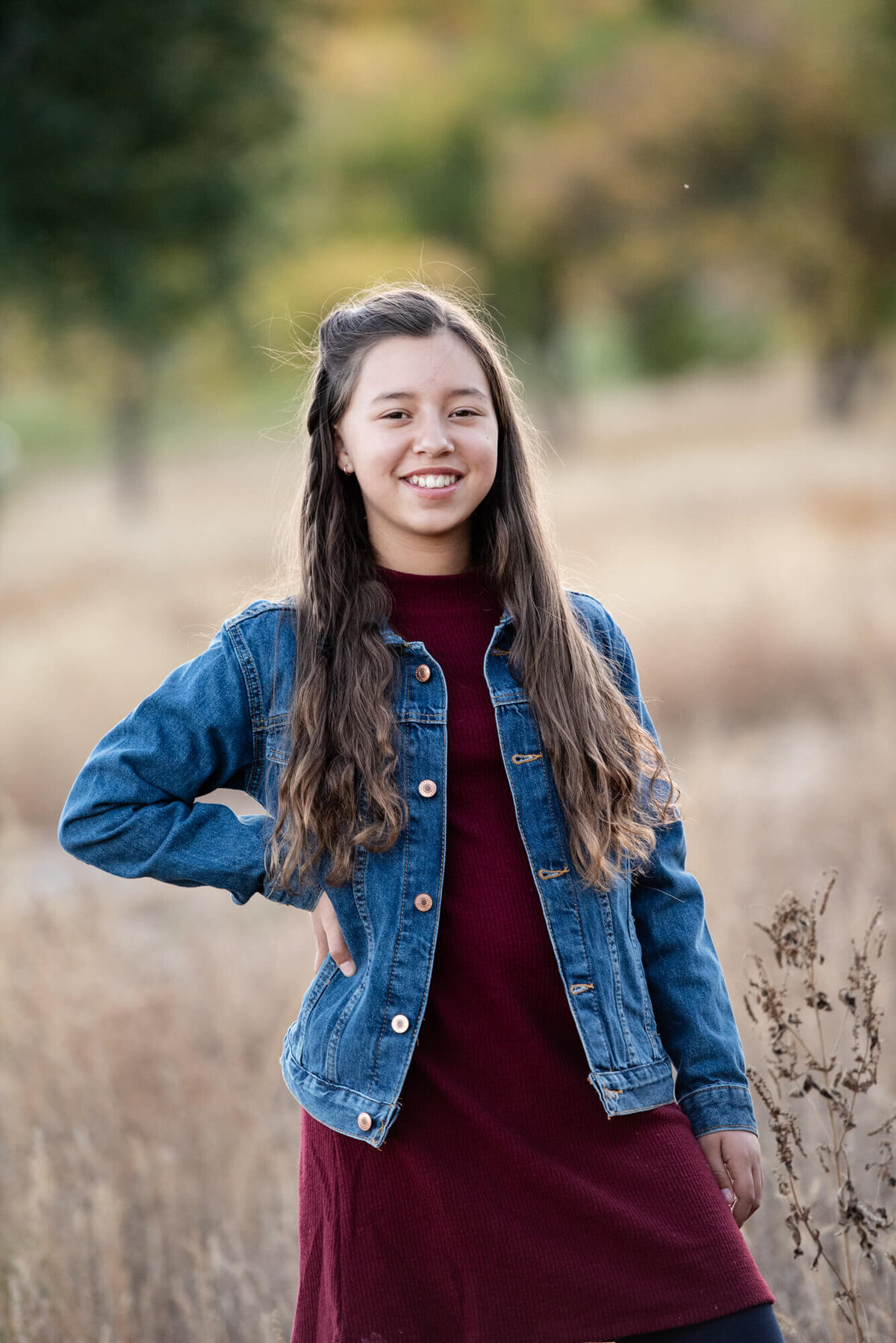 A girl in a denim jacket and purple dress stands in a field with a hand on her hip