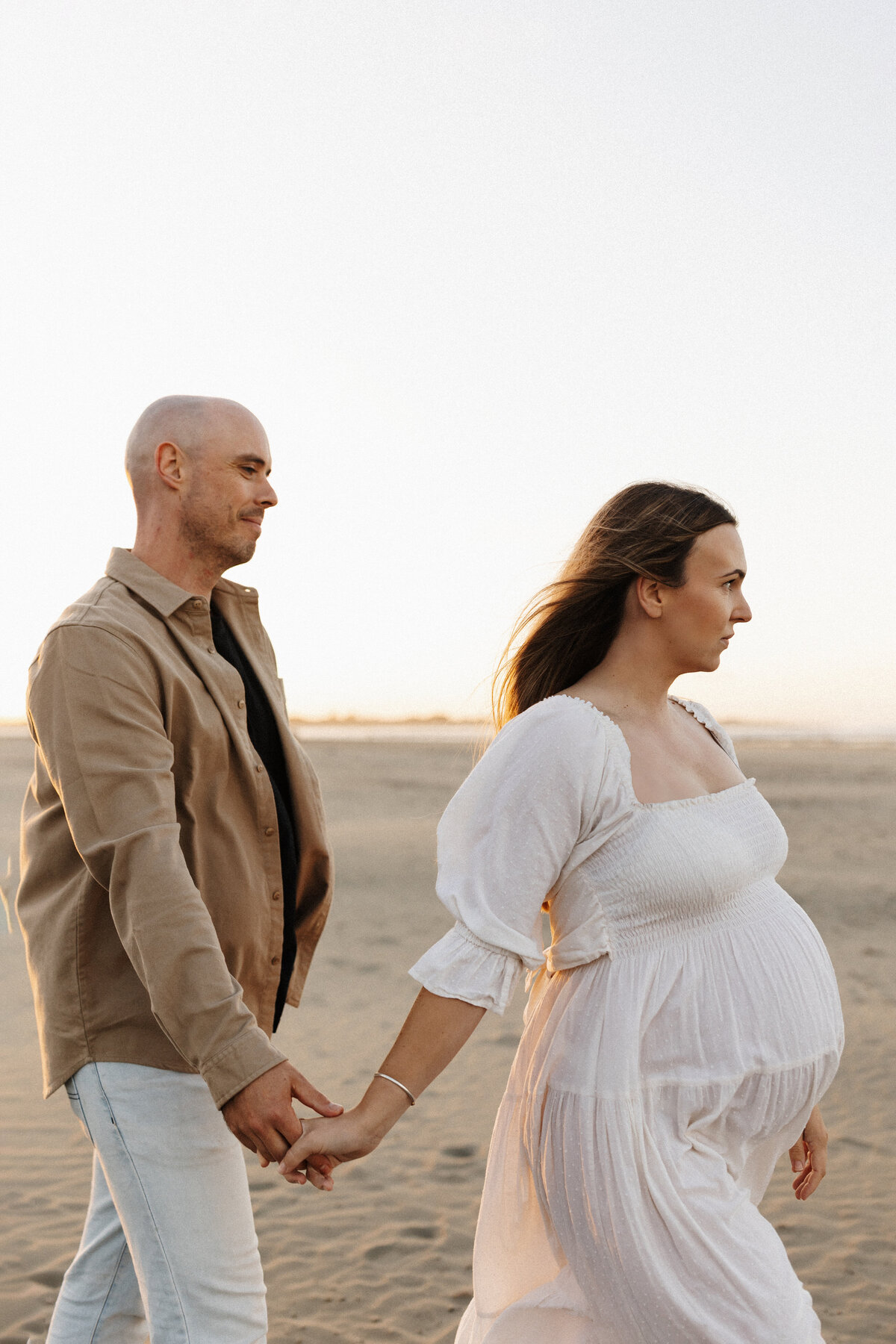 A woman and her husband walk along the beach holding hands during a maternity photoshoot while lucy jane takes candid photographs of them