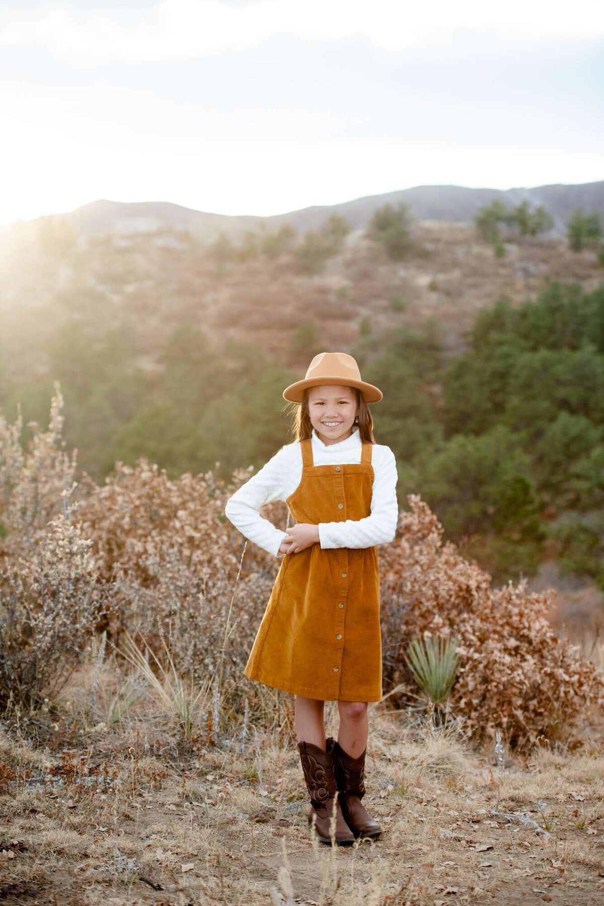 A young girl in an orange dress stands in a trail with hands on her hip and smiling
