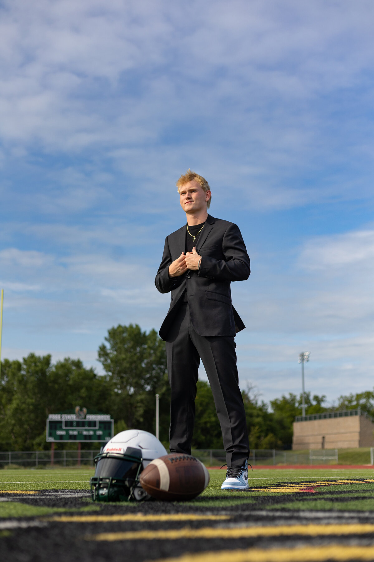 A young man dressed in a black suit standing confidently on a football field with a football helmet and gear beside him.