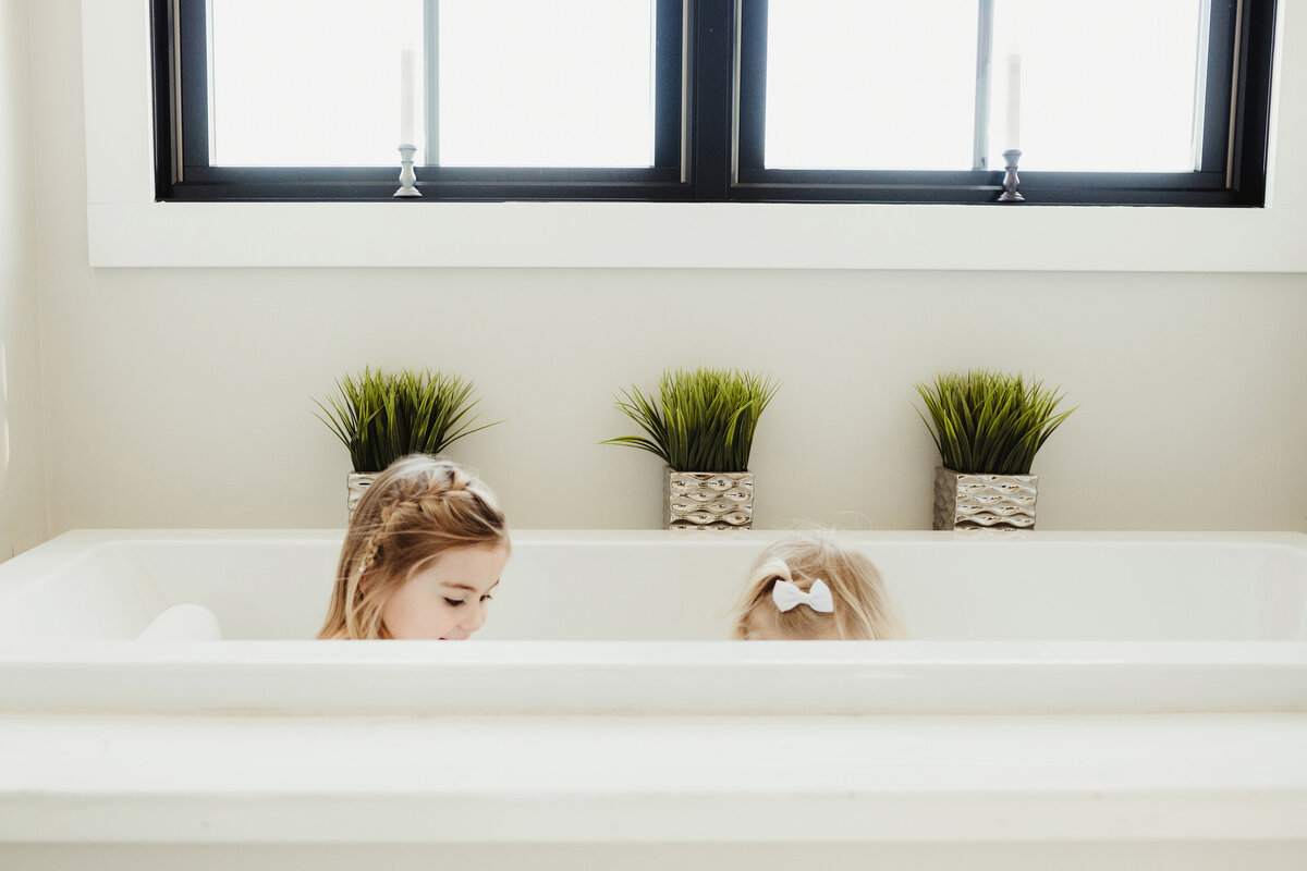 Two little girls are playing in a big white bathtub in natural light.