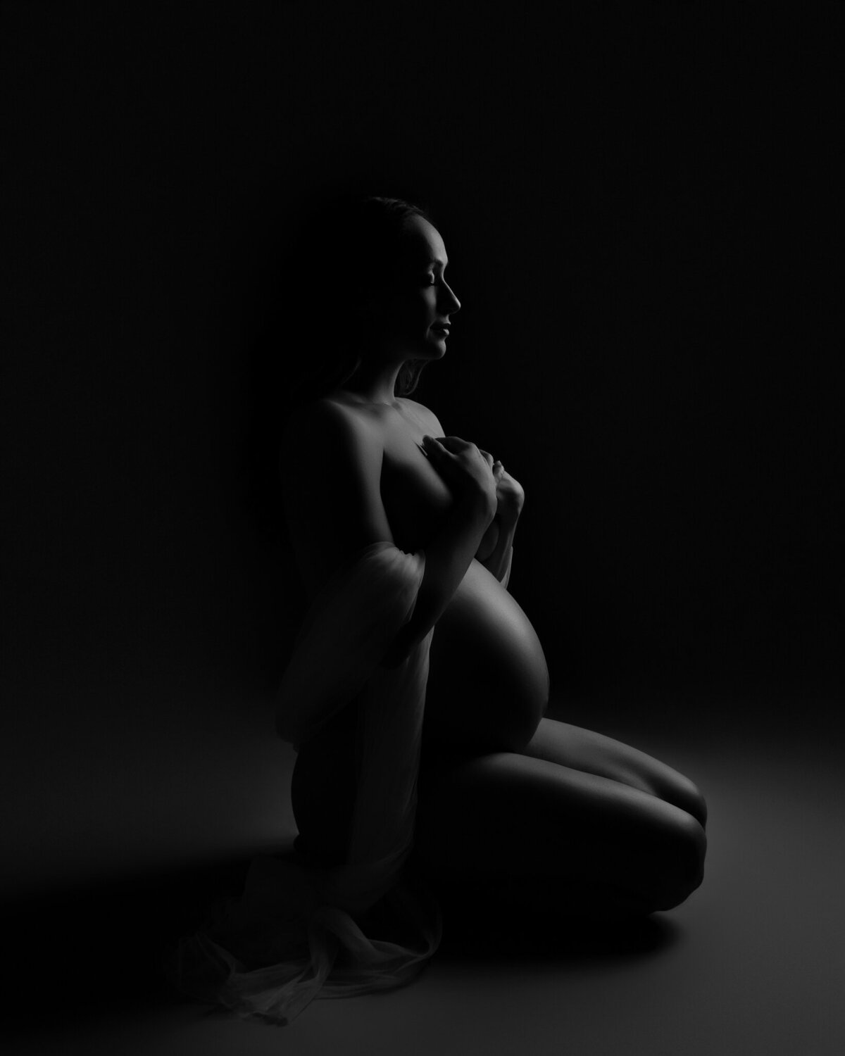 Black and white artistic maternity portrait of a pregnant woman kneeling with sheer fabric, taken in Liberty Hill, TX.