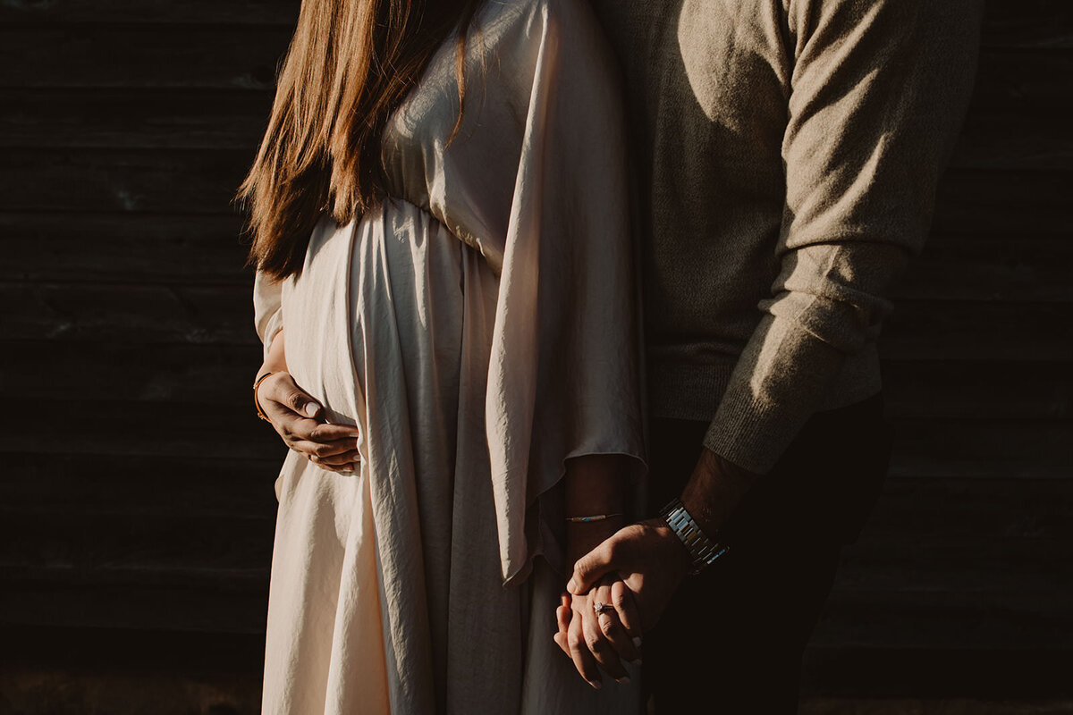 A person standing behind their pregnant partner holding the bottom of their stomach 