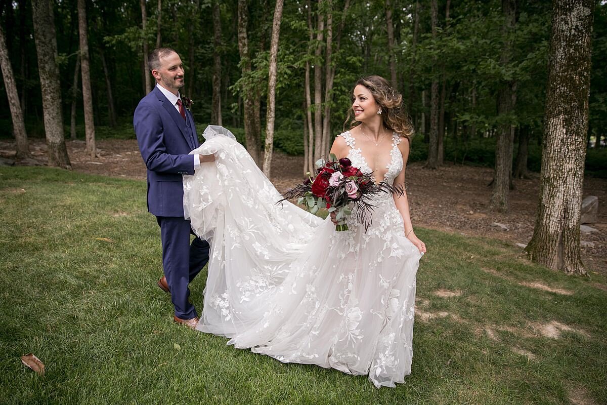 Groom dressed in a navy blue suit holds the bride's long lace wedding dress train as she walks ahead of him. The bride is wearing a lace wedding gown with a deep plunging neckline as she holds a large burgundy and blush bouquet of roses, ranunculus and peonies.