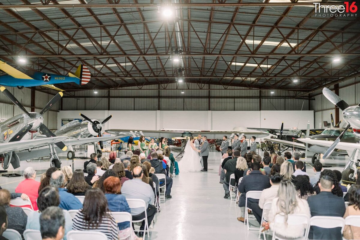 Bride and Groom face each other at the altar as guests look on at the Planes of Fame Museum wedding venue inside a hangar