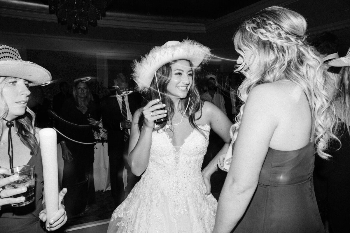 Bride in a white dress with a feathered hat smiling and talking with guests at a lively party.