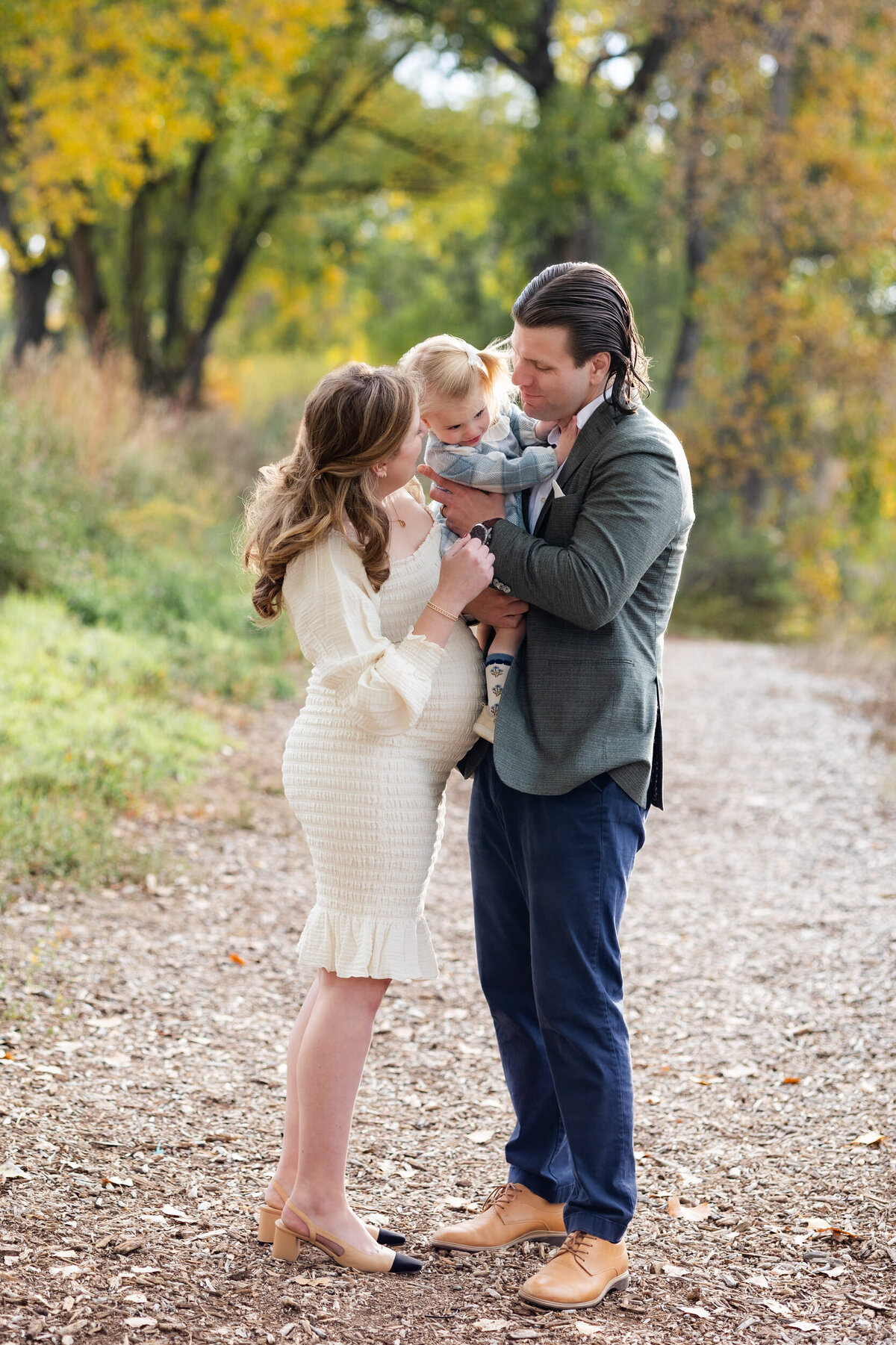 Mom and dad hold toddler daughter between them and smile at her.