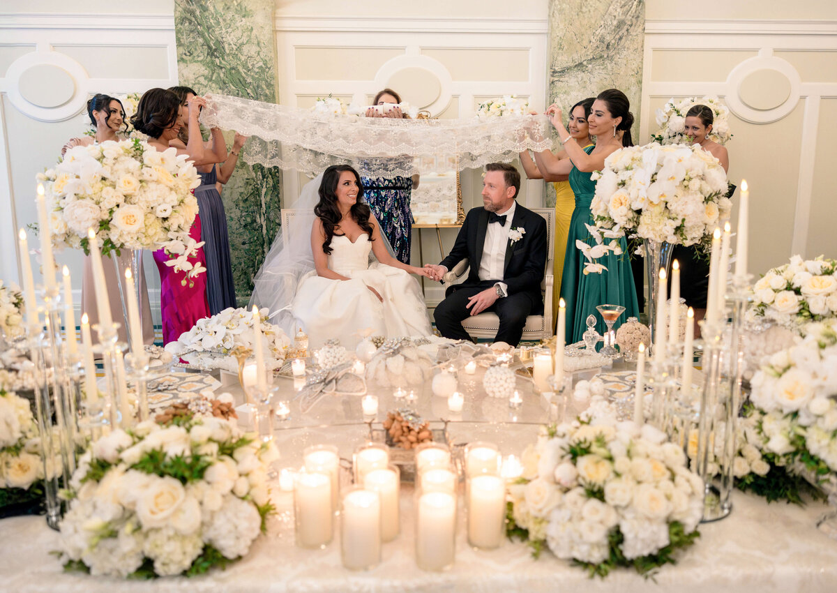 A bride and groom sit together smiling, holding hands. They are surrounded by floral arrangements and lit candles. Several women in colorful dresses hold a lace cloth above them, creating a decorative and festive atmosphere.