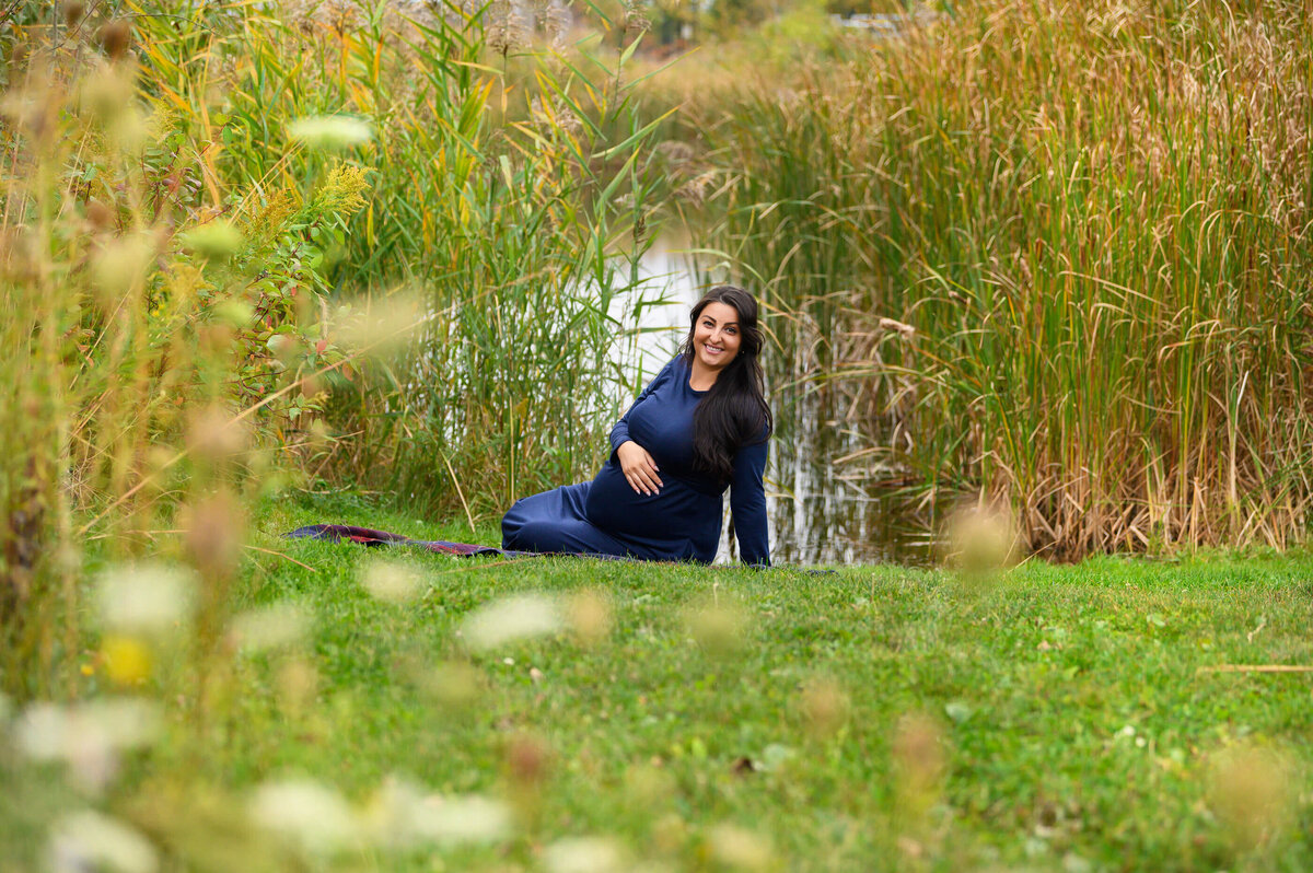 pregnant woman sitting on the ground in front of a pond wearing a blue maternity dress for her fall outdoor maternity photos in Burlington, ON