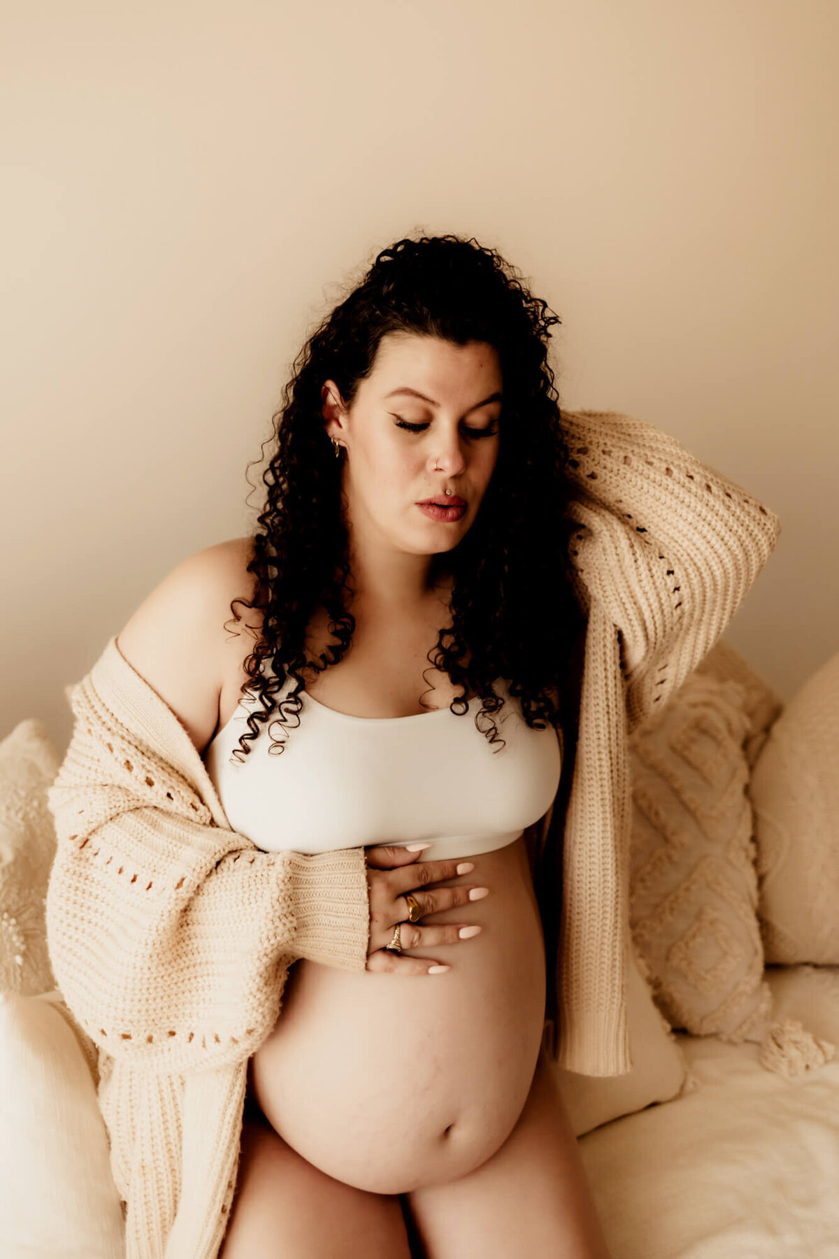 Pregnant woman wearing a bralette and beige sweater holds her hand in her hair while placing the other hand on her belly.