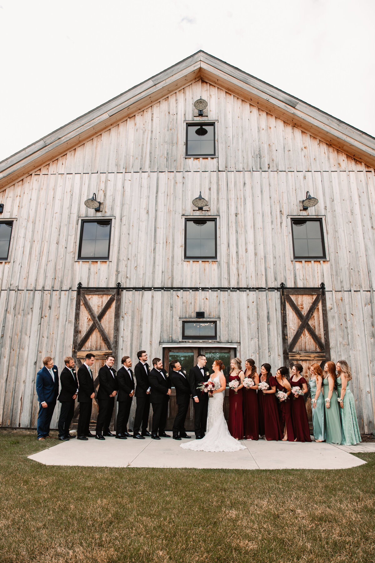 Newlyweds stand looking at each other with their wedding party surrounding them at the entrance to their barn venue