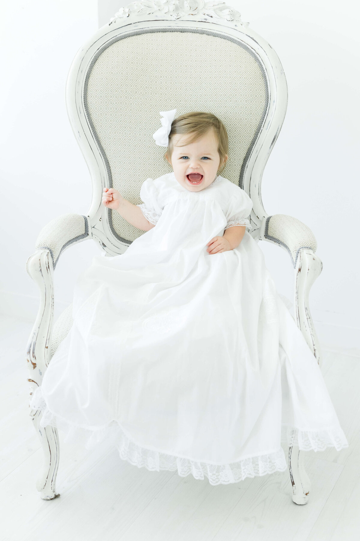 Baby girl in white dress with bow sitting up in chair for milestone photography