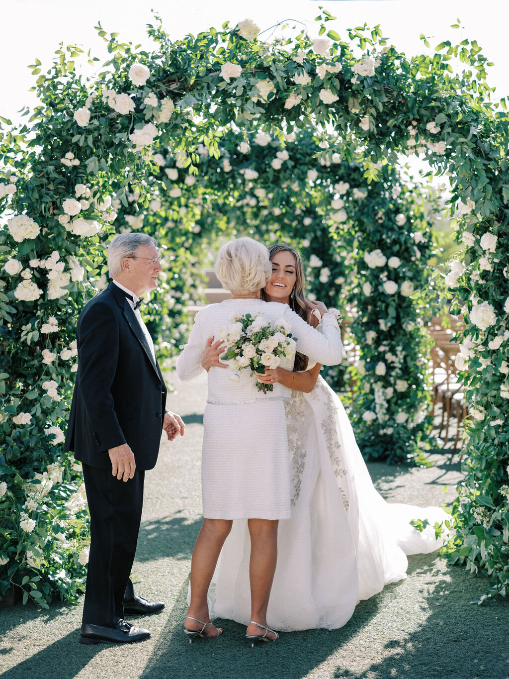 The bride shares a heartfelt embrace with family under a gorgeous floral arch, capturing a meaningful wedding day moment.