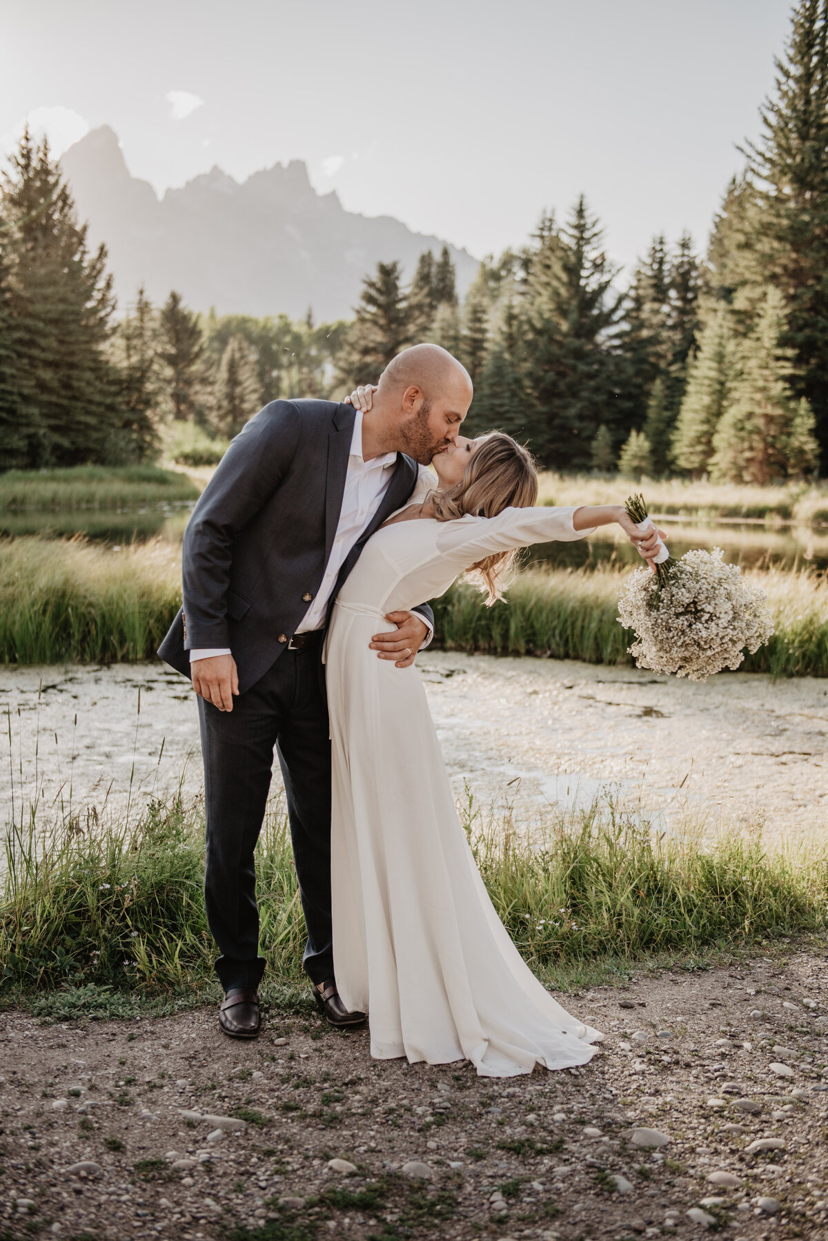 wedding couple standing in front of a river in the Tetons kissing with the man's hand is holding the woman's waist and the woman is bending back with her arm in the air with her wedding bouquet