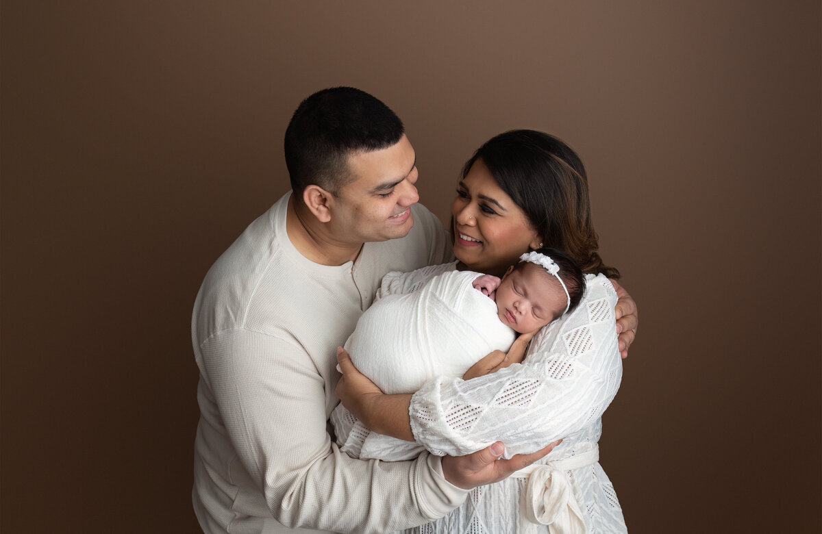 A warm and intimate family portrait captured in-studio, featuring both parents holding their newborn daughter swaddled in white. Photographed at Meraki Photographic Creations in Paris, Ontario.