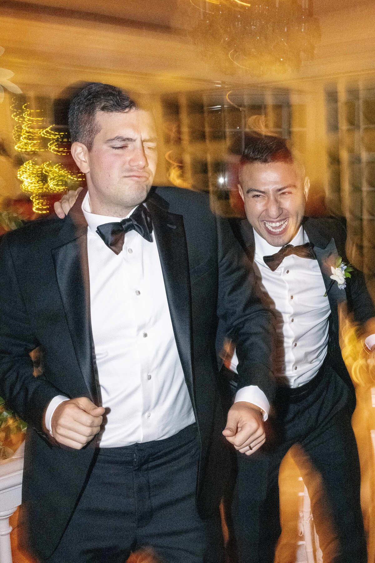 Two men in tuxedos are joyfully dancing at an event. One man has his eyes closed, and both are smiling broadly. They are holding arms while surrounded by blurred lights, suggesting movement and a festive atmosphere.