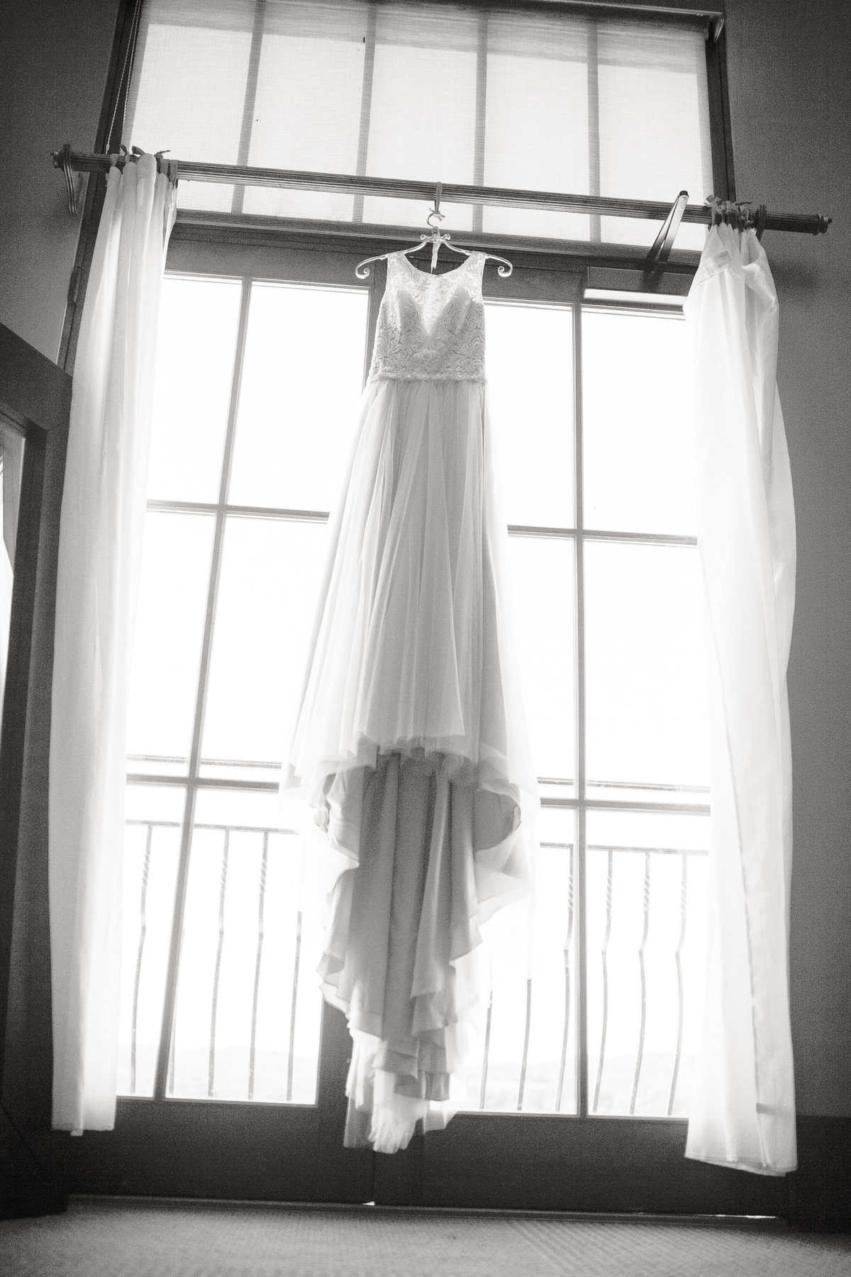 B&W image of wedding dress hanging in the window of the bride's home.