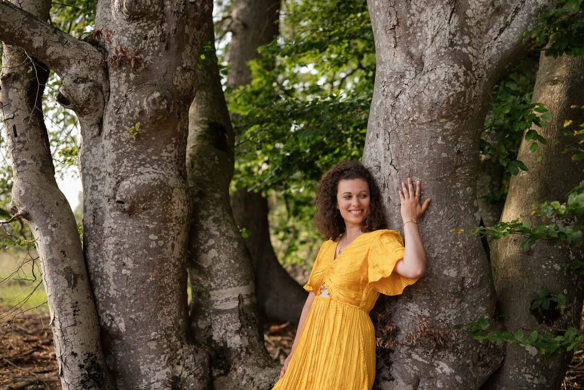 eine Frau in gelben Kleid steht im Wald an einen Baum gelehnt und ist mit der Natur verbunden