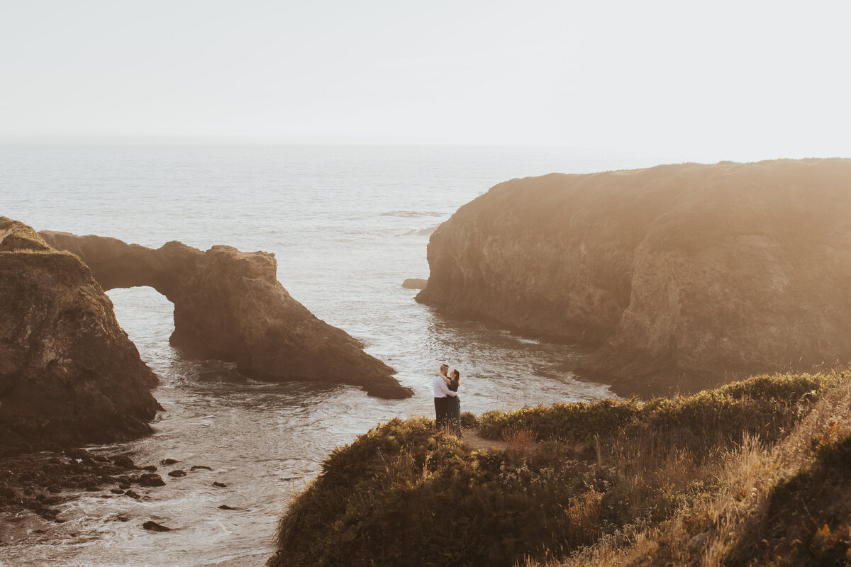 A couple stands on a coastal cliff at sunset, framed by breathtaking ocean views, highlighting editorial wedding photography.