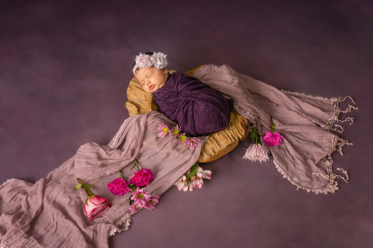 newborn baby girl in a bowl on a purple background.  There are pink flowers and the baby is wrapped in plum purple.  She is wearing a purple floral headband and is laying on her back.