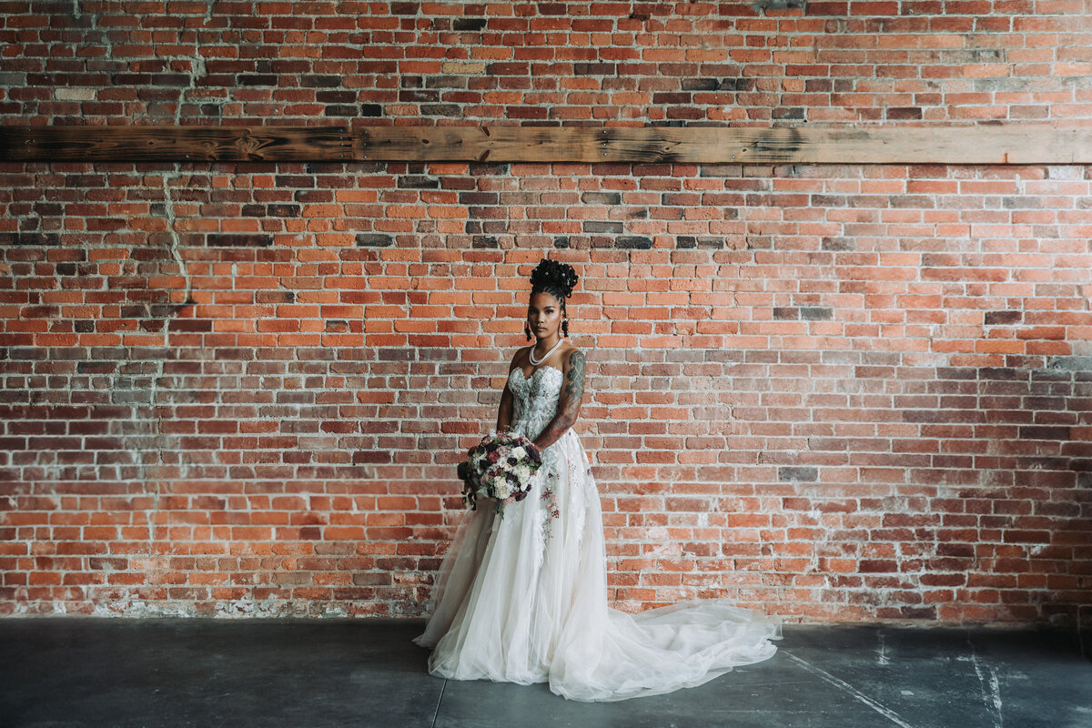 Bride stands with bouquet against an exposed red brick wall for a dramatic wedding portrait at The Neon House in Norfolk, Virginia.