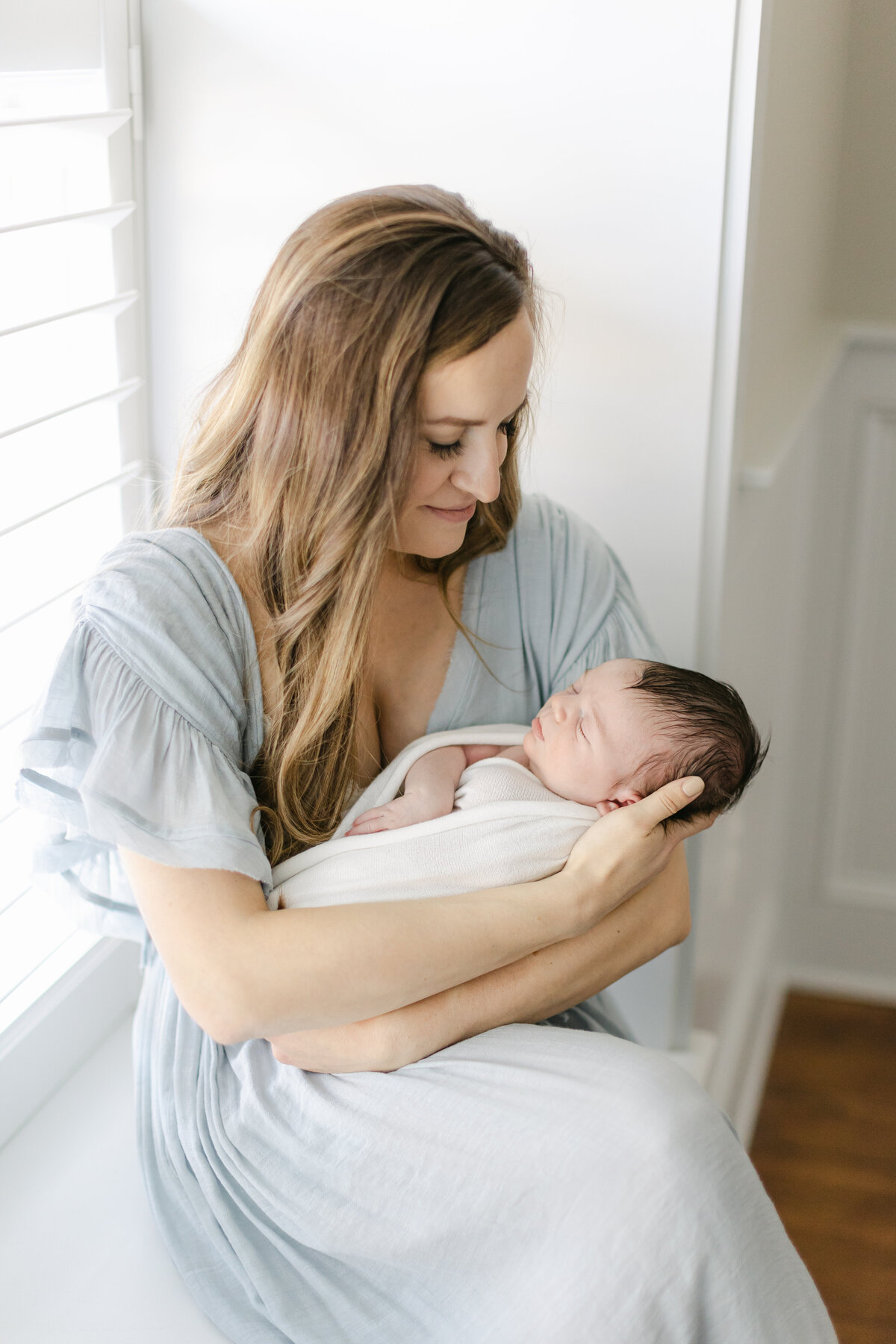 mom wearing a blue dress sitting on the edge of a window seat holding her infant baby with loving eyes photographed by Philadelphia Newborn Photographer Tara Federico