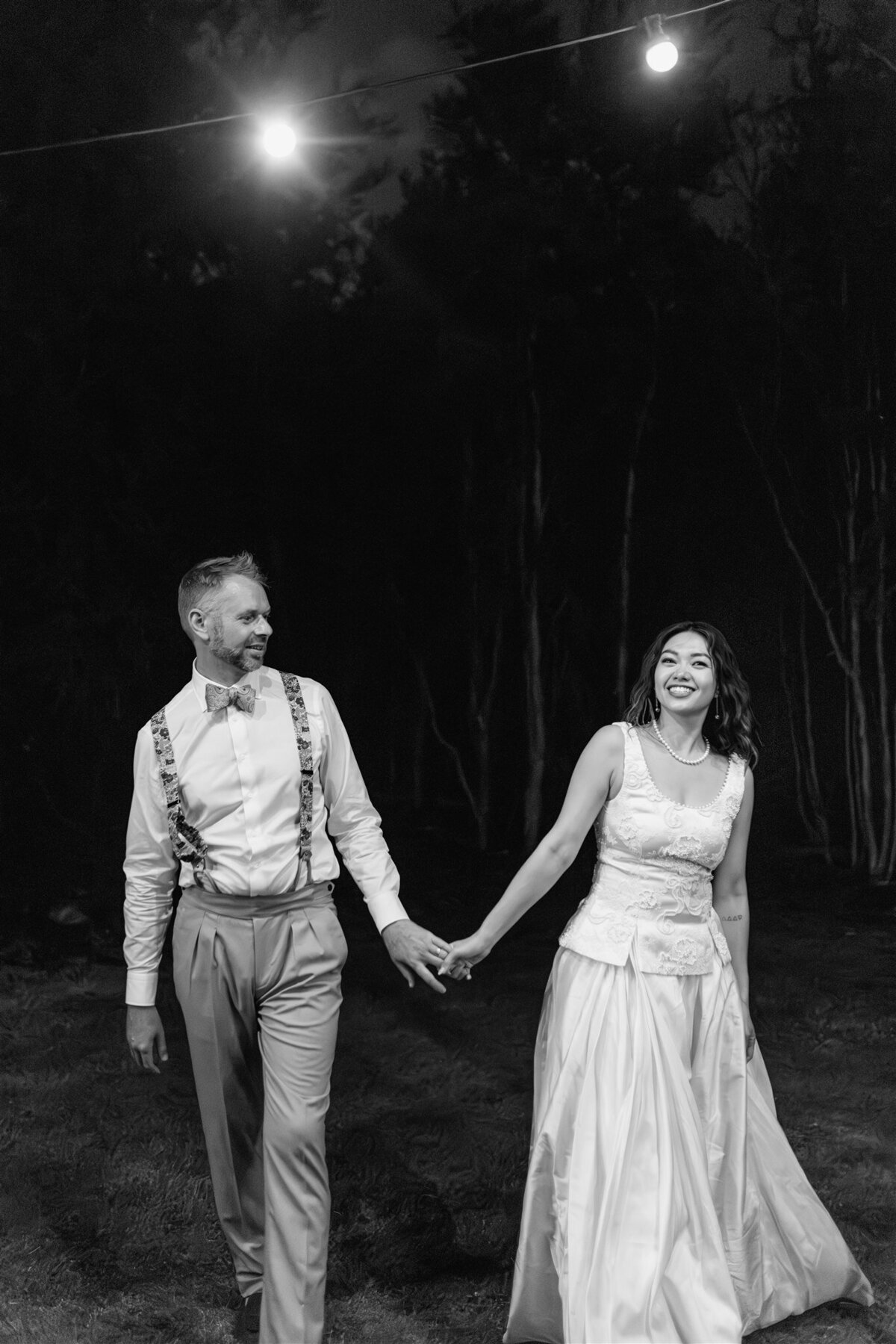 A bride and groom walk hand in hand in the dark, smiling at each other under twinkling festoon lights, captured in black and white.