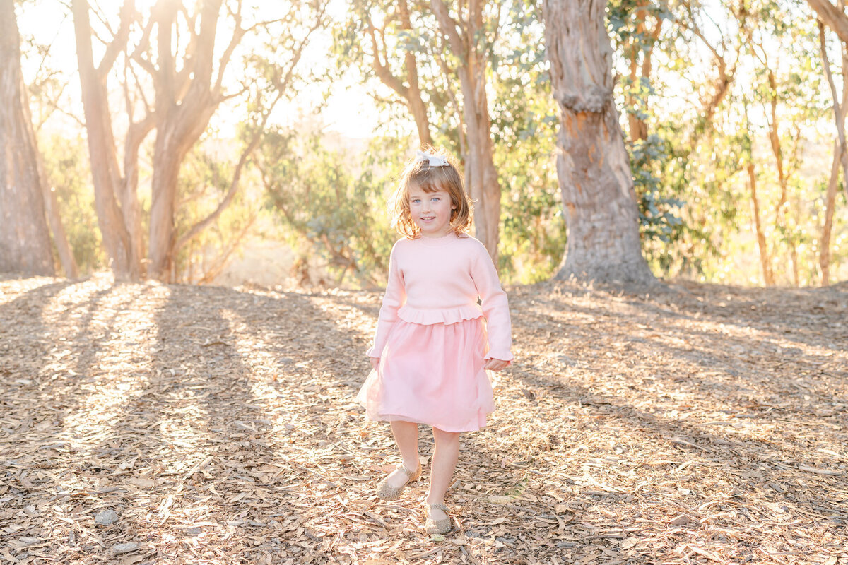 little girl in a pink dress walking towards the camera in front of a grove of eucalyptus trees in the san francisco bay area