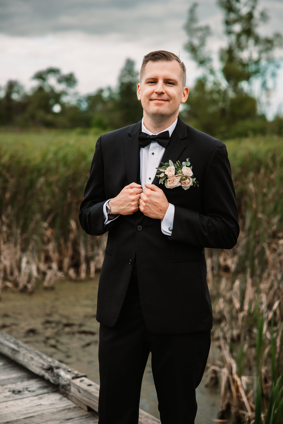 A groom in a black tux stands on a bridge over a pond holding his lapels
