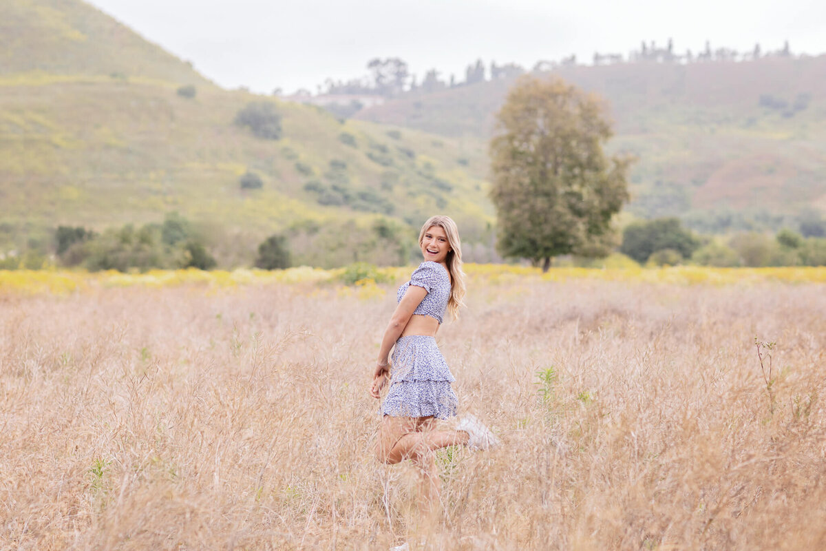 A woman in a light blue dress stands in a field of tall grass, looking back with a smile. The background features rolling green hills and a single tree under a cloudy sky.