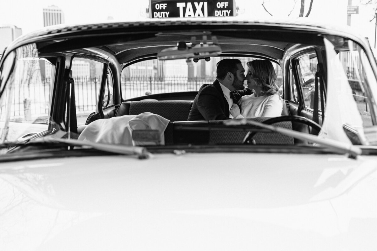 Black and white photo of the bride and groom kissing inside of a taxi. Shot from the outside.
