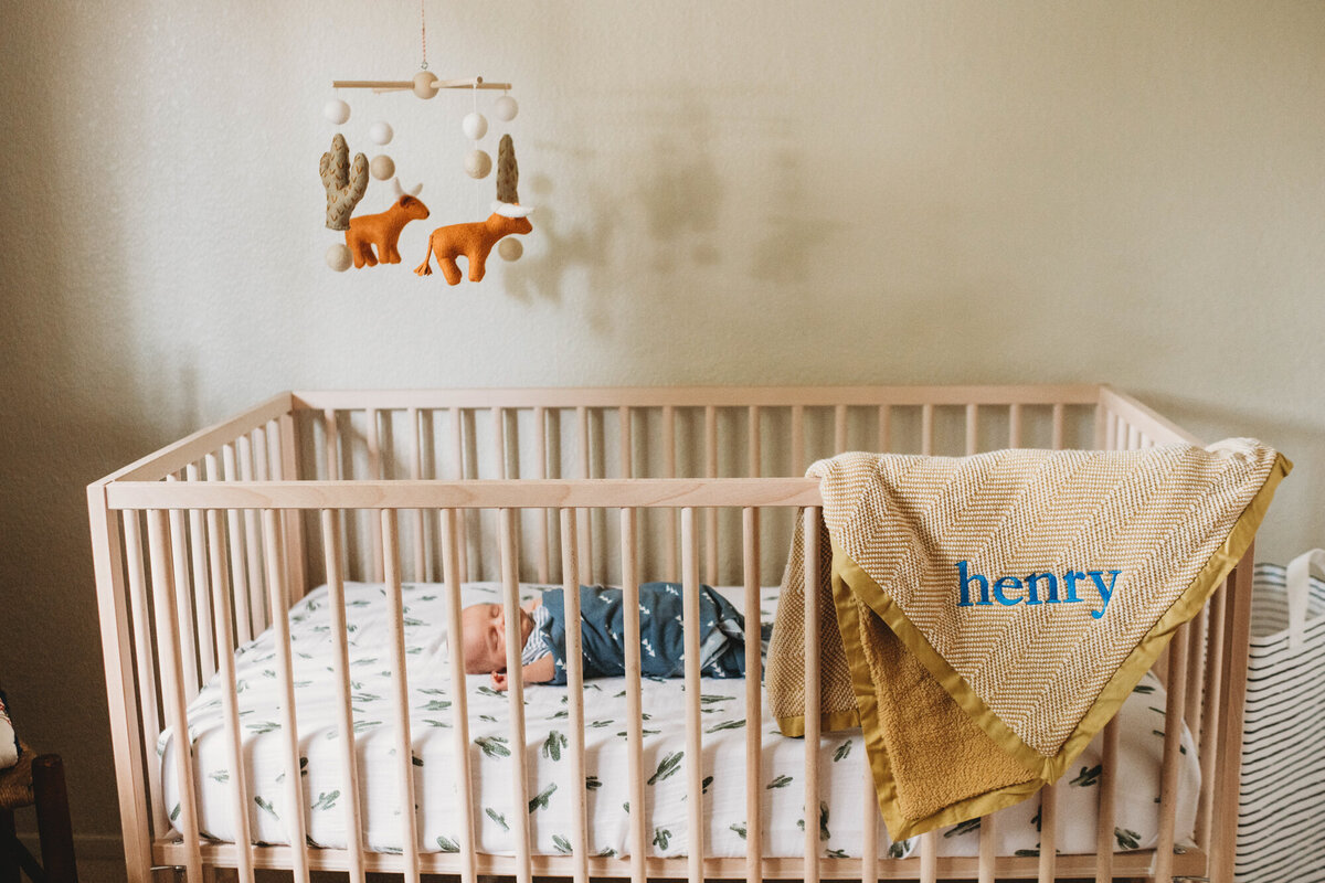 Newborn Photographer, Baby boy sleeping in a crib with a blanket with the name "Henry" draped across the crib railing.