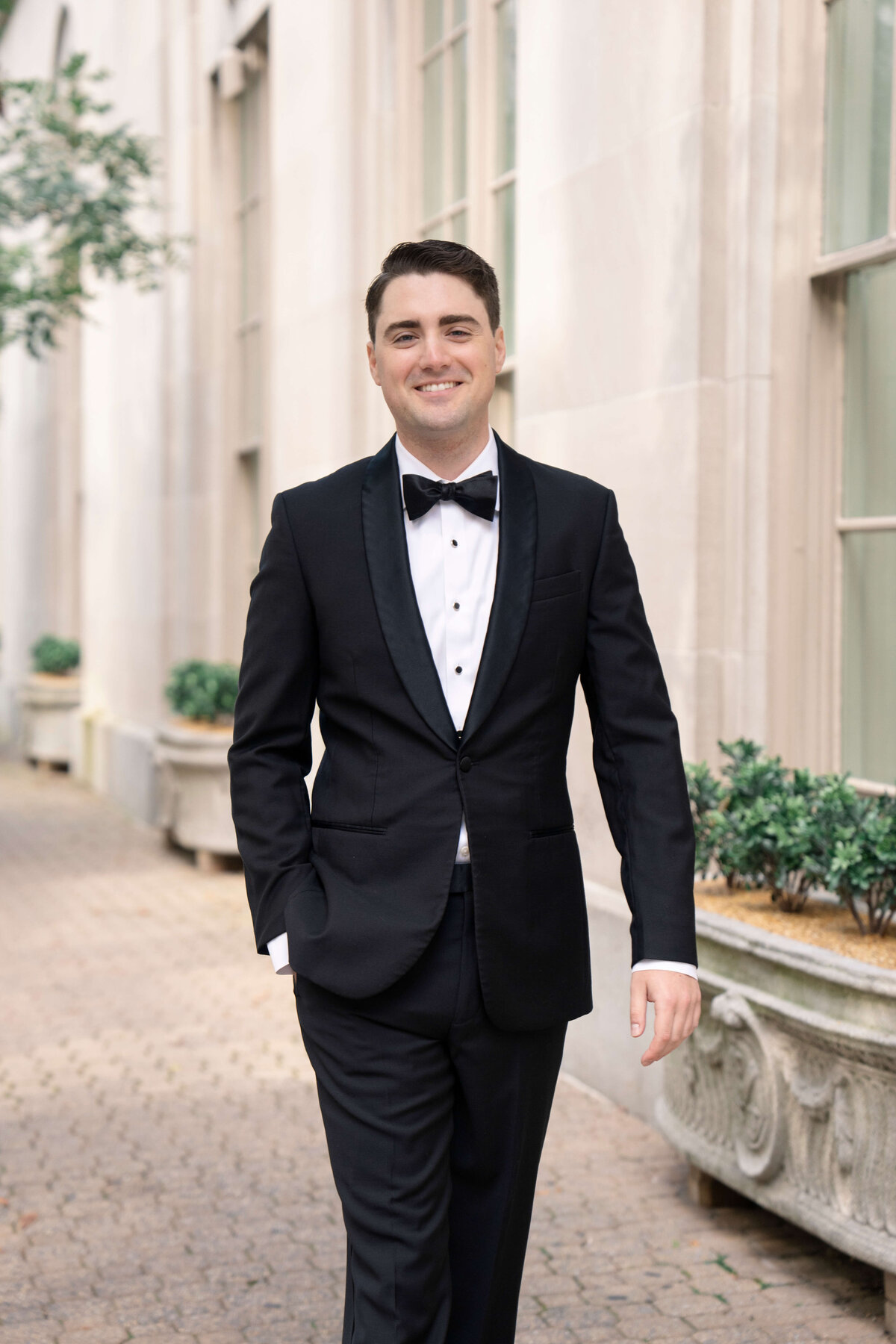 A man in a black tuxedo with a bow tie is walking outdoors on a paved pathway. He is smiling, with one hand in his pocket. Behind him are large windows and greenery in rectangular planters attached to a white building.
