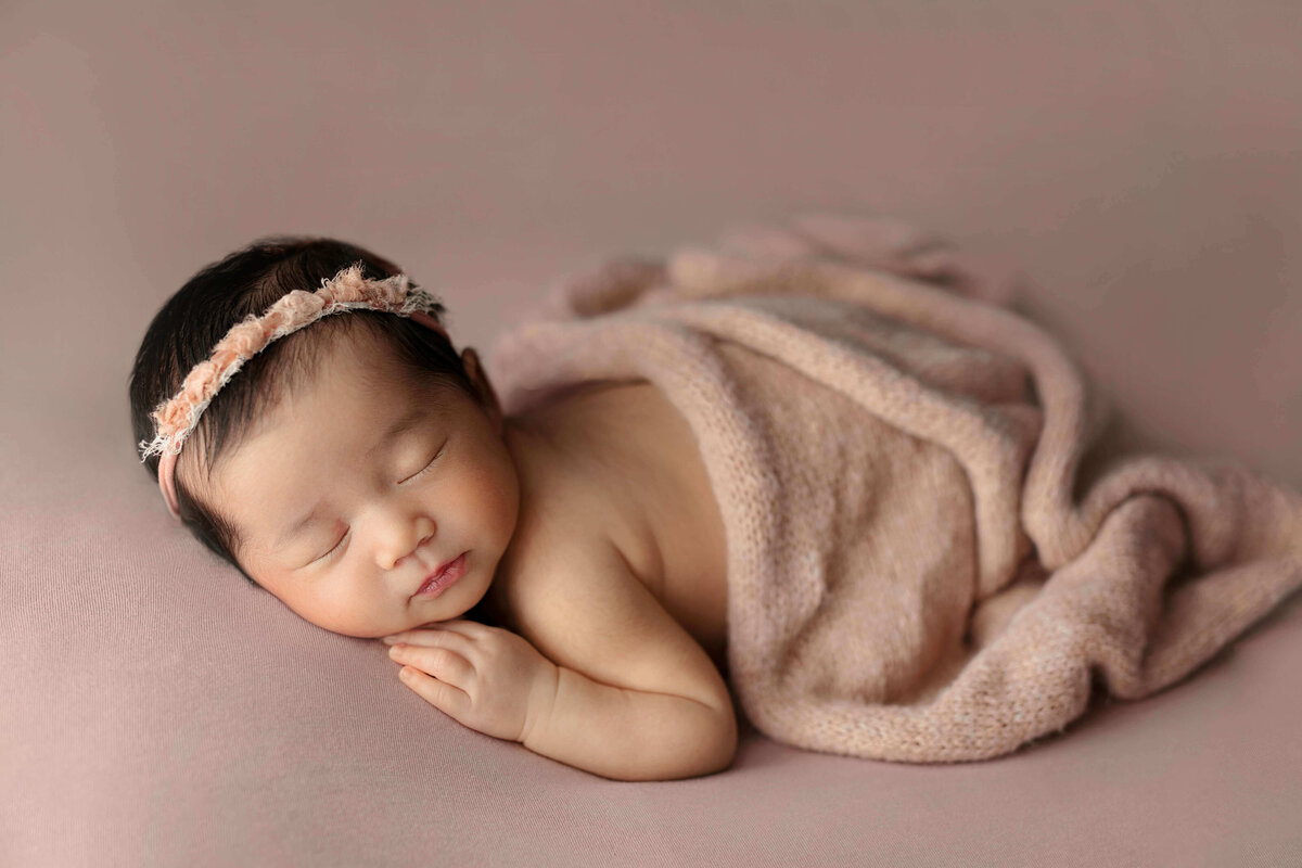 Newborn girl laying on her tummy  on a pink backdrop with matching headband  and wrap at a baby photo shoot in Northern VA