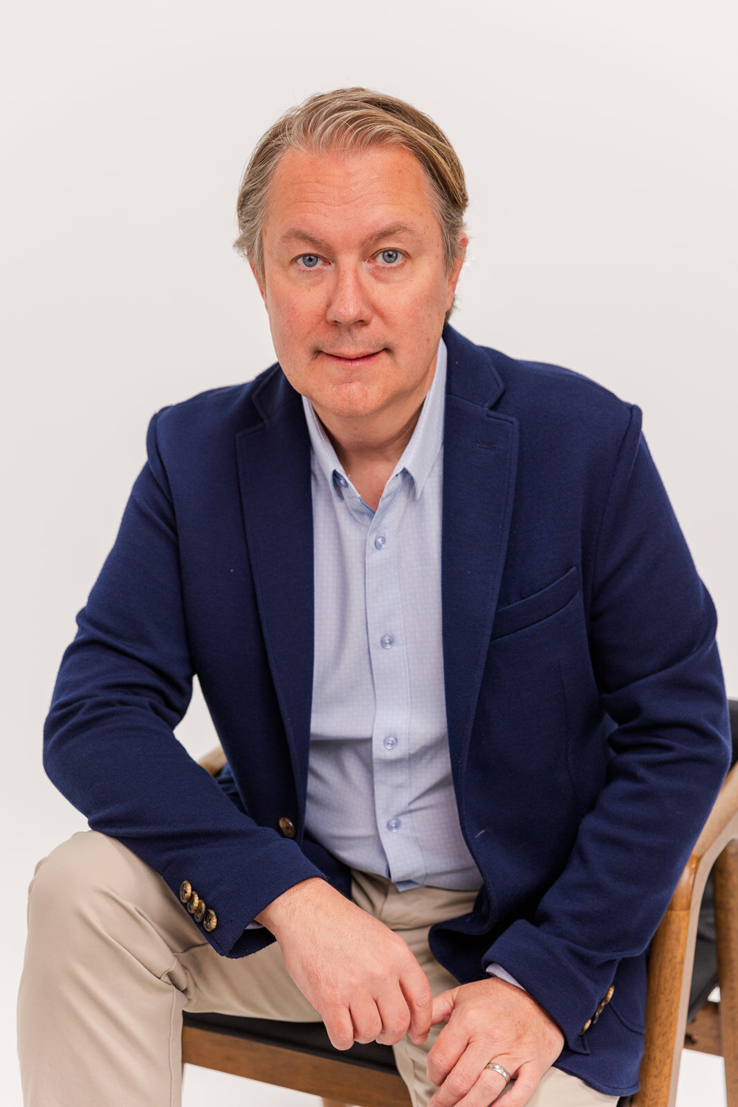 man wearing a kaki pant, blue shirt and blue jacket sitting on a chair in front of a white backdrop during portraits session