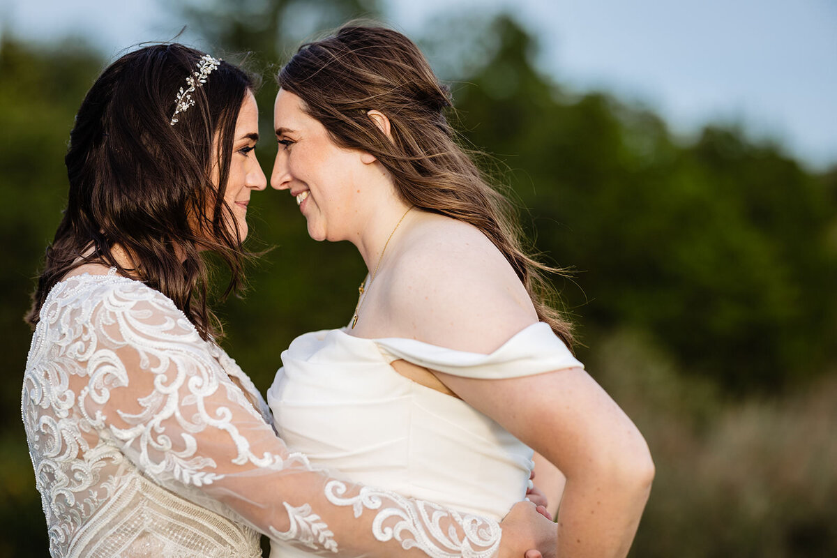 A close-up of the two brides facing each other, foreheads touching, one with a lace dress and the other in a simple white dress, both smiling.