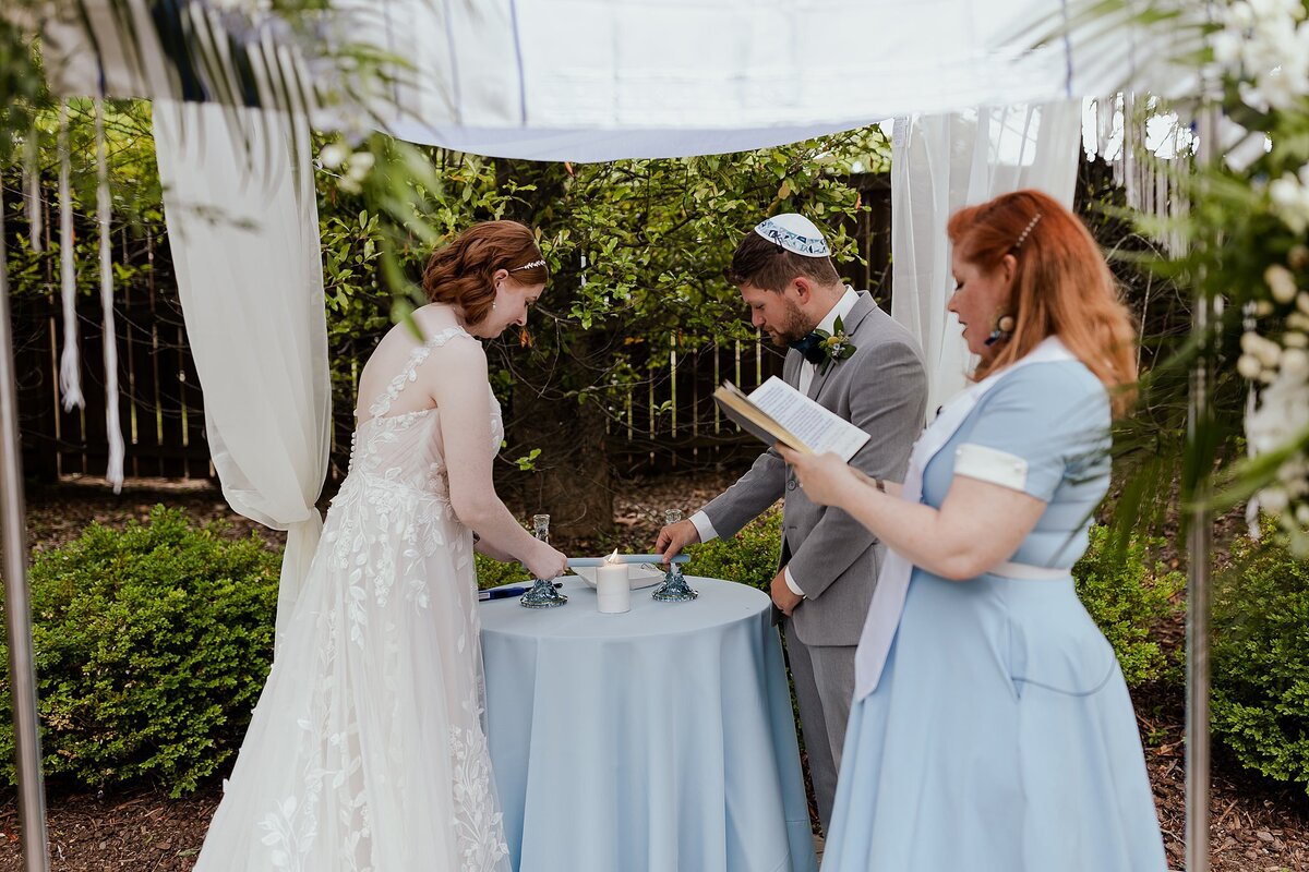 redheaded bride wearing a floral lace wedding dress stands under the chuppah with the groom wearing a white yarmulke and light grey suit. The redhaired officiant stands to the side as the bride and groom light the unity candle.