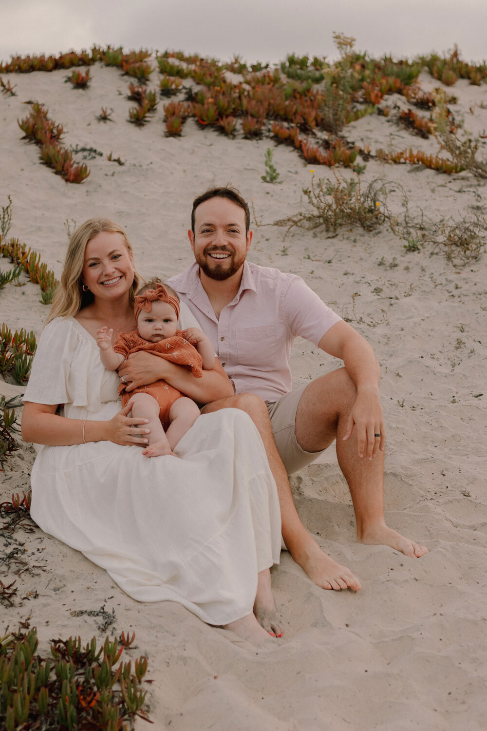 Family sitting on sand, smiling with baby in the center, surrounded by greenery