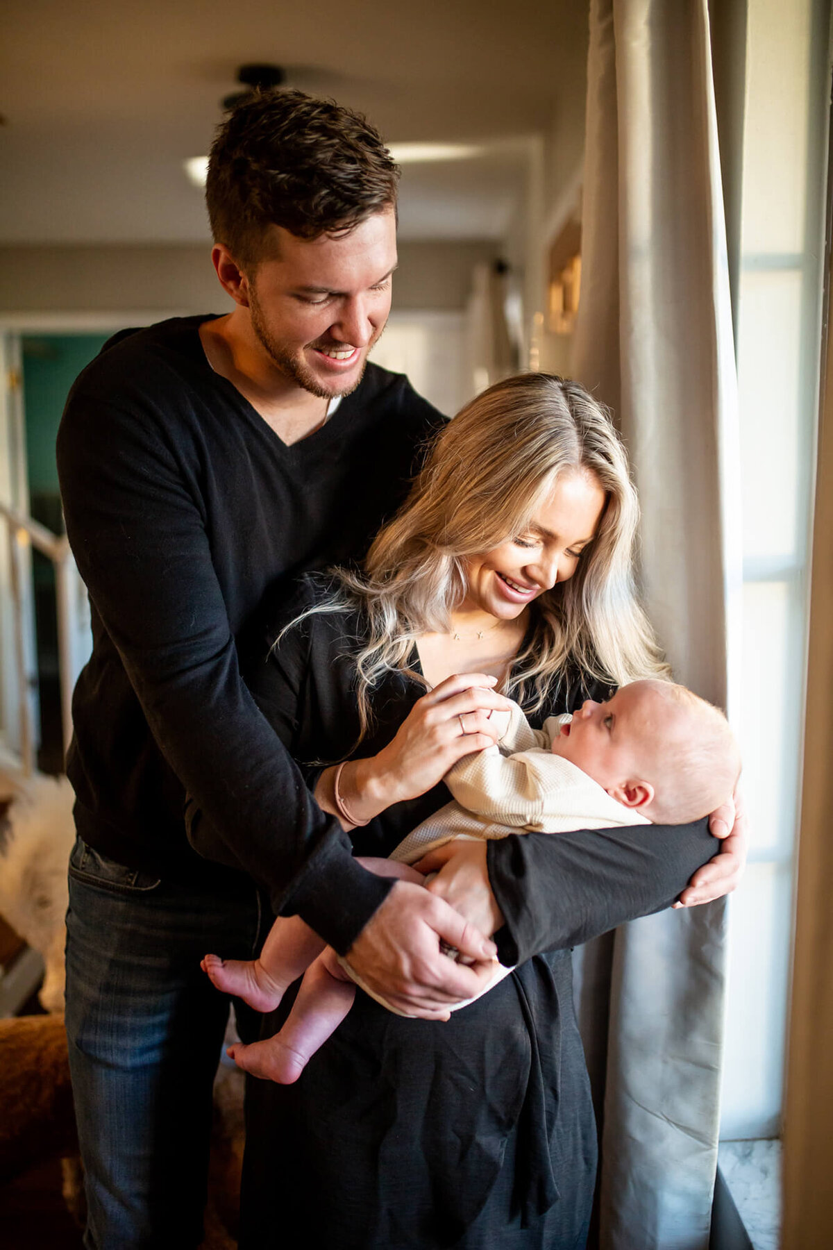 In home natural Family session. Mother is cuddling baby by window light while father has his arms wrapped around both of them. Located in Riverview, FL 