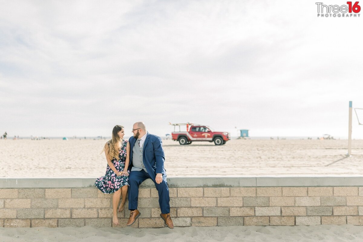 Newly engaged couple sit together and gaze into each other's eyes while sitting on a brick wall at Huntington Beach